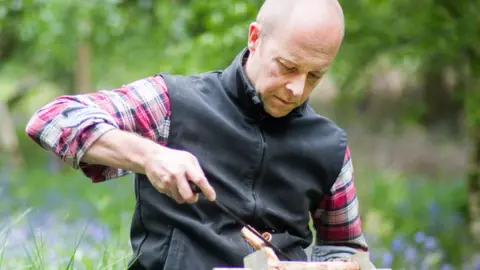 Clare Bowes Furniture maker Nick James working with wood in an outdoor forested area. He is wearing a red white and blue check shirt and black gilet. Seated, he is carving a branch with woodworking tools.