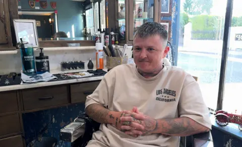 Kieran Eljay Eaton sits in a barber chair with a window behind him. He has his hands folded in front of him and is wearing a white t-shirt with the words Los Angeles, California, on front top left. He has tattoos on his arms and hands and a short back and sides haircut