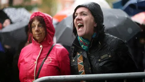 Reuters A woman in a black raincoat shouts over a barrier as another woman in a pink raincoat looks on. There are several umbrellas behind them.