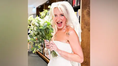 A woman with long blonde hair and wearing a white strapless wedding dress and white veil is smiling while holding a bouquet of flowers. She is standing in front of shelves of books as part of the Storyhouse library.