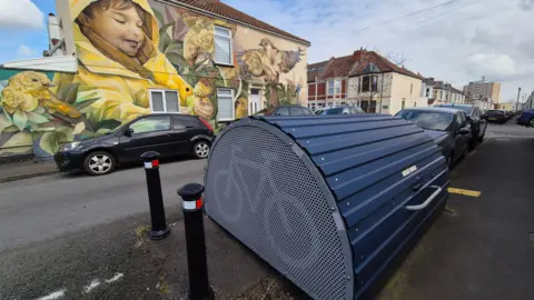 A bike hangar installed on a residential road in Bristol. It is a blue corrugated item with a curved structure and has bollards either side. In the background is a house with a large colourful mural painted on the side