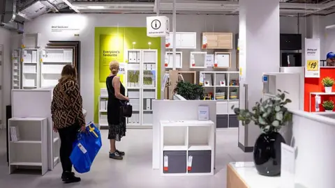 Bloomberg via Getty Images Two women, one holding a large blue plastic bag, stand in an Ikea store looking at white-coloured shelving and storage units