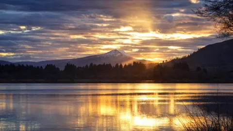 A snow-capped Ben Lomond in the distance, with Lake Menteith in the foreground, the sun is reflected on the rippled water.