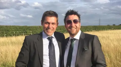 Jonathan Rudd Jonathan and George Rudd wearing dark suits, light shirts and ties, smiling towards the camera, standing in front of a field of hay grass with a long hedgerow behind them