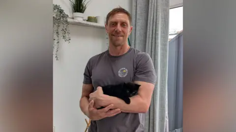 A man with a grey T-shirt holding a tiny black kitten in his arms. He is standing inside a house with a curtain in the background.