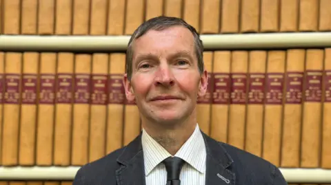 BBC Dr Alan Borthwick has short dark hair and is wearing a  dark jacket, white shirt and dark tie. He is standing in front of two shelves of official records books