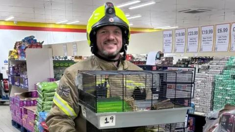 Essex Fire Service A firefighter wearing a yellow hi-vis helmet holds a black crate inside the pet shop. Lots of stock is stacked in rows behind him, most of which are bags or tins of pet food.