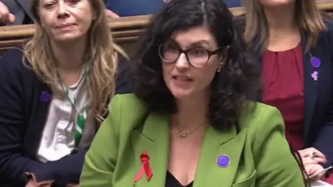Layla Moran delivering her PMQ in parliament. She wears a green jacket, with a red ribbon on it and a purple broach. 