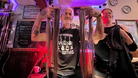 BBC Rick presenting his cut off grey hair to the camera in a pub lit up with red lighting.