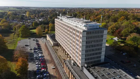 A drone image of a large office building in a rectangular shape, with trees, a car park and housing around it.