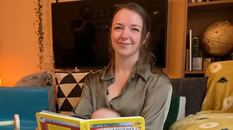 A woman reads a book called Hairy Maclary to her child. The book is obscuring her son’s face. They are in a living room.