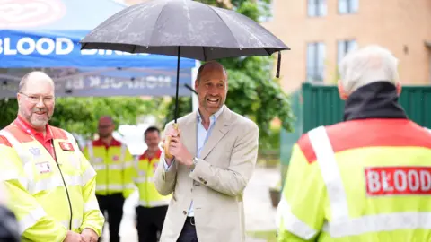 PA Media Prince William, dressed in dark trousers, a pale jacket and pale shirt, holds a black umbrella as he talks to men in hi-vis jackets. One has "BLOOD" written across the back of his hi-vis 
