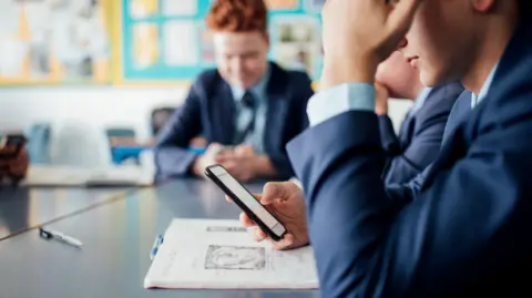 A child in a school uniform sat at a desk holding a smartphone.