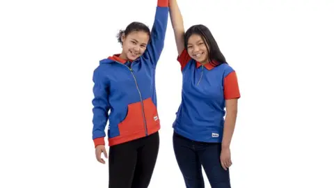 PA Media Two girls stand next to one another, they are holding hands in the air. The girl on the left has dark hair tied back and is wearing a blue and red hoodie with a zip down the middle and black leggings. The girl on the right has long, dark hair and is wearing a red and blue polo shirt with a three-quarter zip, and jeans. The background behind them is plain white.