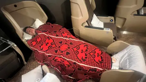 A man tries out a fully reclining pod on a ferry, he has dark hair and is covered in a red and black patterned blanket