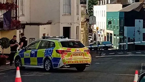 A narrow street with white walled buildings on both sides and a police car parked in the foreground with blue and white police tape blocking the street