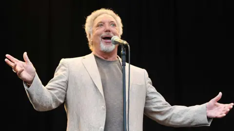 Andy Sheppard/Redferns Tom Jones performing onstage singing into a microphone. 