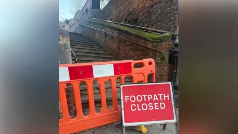 Shropshire Council An orange barrier with red and white stripes on it is blocking a set of steps running alongside a brown brick wall. Also in front of the steps is a red sign with white writing that reads "footpath closed"