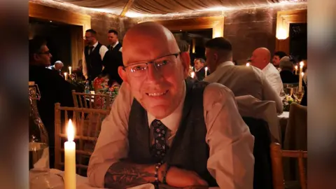 Darren Burke Mr Burke in a white shirt and dark waistcoat smiling at the camera with his arms crossed on the table in front of him. He Has dark glasses and is sitting down at an event, with a candle burning on the table just in front of him. 