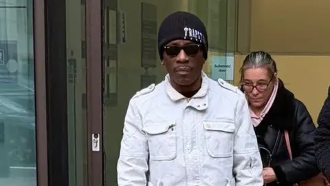 Henry Morgan, a man wearing a black woollen hat, pale denim jacket and sunglasses, pictured close-up exiting a set of glass doors.