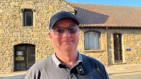 A man wearing a black cap and tinted glasses smiling looking into the camera. He's wearing a navy polo top. There's an old stone building behind him.