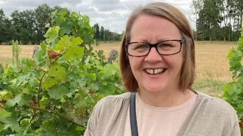 Woman smiles at the camera,  wearing glasses with green fields in the background