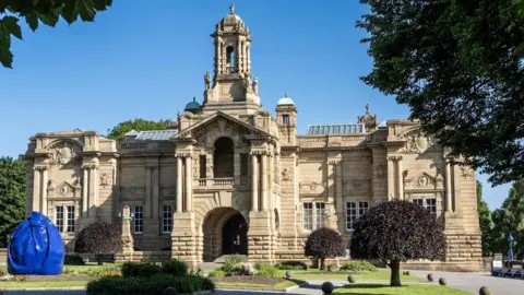 Bradford Council Bradford’s Cartwright Hall Art Gallery: A large, stone building set within landscaped grounds. In the foreground, there is a garden with trimmed grass and flowerbeds. 