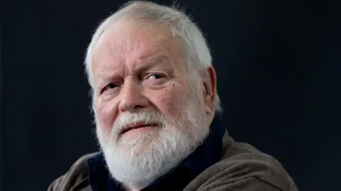 Michael Longley has short white hair and beard and is wearing a brown cardigan over a dark blue shirt.