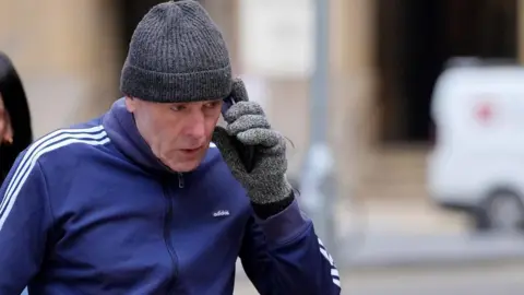 David Chick, a man wearing a grey knitted hat and grey gloves, and a blue and white tracksuit, holding a phone as he arrives at Southwark Crown Court