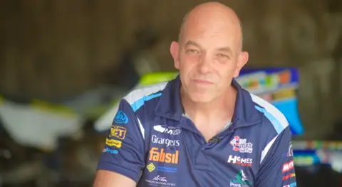 Jamie Niblock/BBC Simon Huntingford in his blue team shirt, emblazoned with sponsors. He is bald and looking onto the camera.