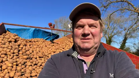 A man stands in front of a large pile of brown, unwashed potatoes, stacked on a red trailer under a blue sky. He is wearing a baseball cap, dark-grey fleece and matching polo shirt.