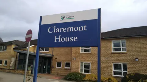 BBC A two storey building with sand coloured bricks and windows with white frames. 
In the foreground is a blue sign with 'Claremont House' the name of the dementia home on. 