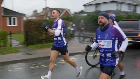 Jamie Niblock/BBC Kevin Sinfield and two other men are running down the street. 