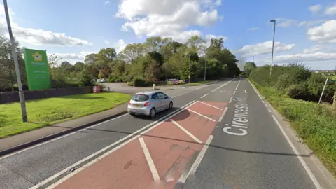 A wide A-road split into two lanes with red hatching in the centre before a right-hand turn. One car is on the road and a garden centre sign can be seen on the left.