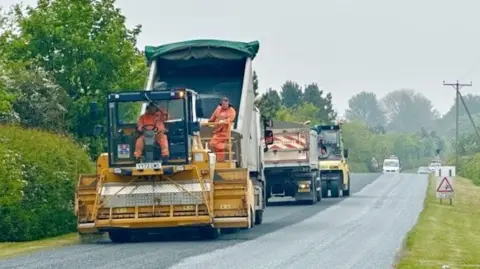 Three large vehicles are laying stone chippings on a long road. There are two men in hi-vis outfits - one is driving, the other is watching the process. There is a 20mph speed restriction sign on one side. 