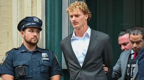 Getty Images Daniel Penny in grey suit with no tie with his arms behind his back, next to police officer in uniform and being escorted by police officer in suit jacket.