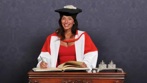 Davina McCall in a graduation gown signing a book