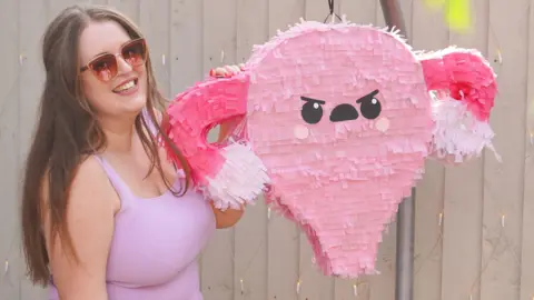 Jen Moore Jen is wearing a pink top and sunglasses, she is standing next to a pink pinata in the shape of a womb and ovaries.