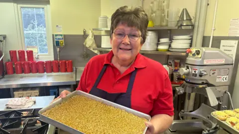 Alison Hopkinson holding a tray of food in a school kitchen. She has short hair, is wearing glasses and a red polo t-shirt and navy apron, and is smiling at the camera.