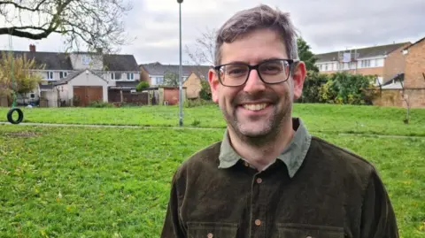 Rob standing in front of a field in a residential area. He is wearing a dark jacket and black rimmed glasses and is smiling at the camera.