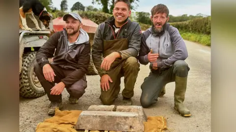 Three men are crouched down on the ground on what appears to be a road. They are smiling with the two ingots placed on the ground in front of them on a mustard-coloured piece of cloth. An agricultural vehicle with a sheepdog perched on it can be seen behind them.