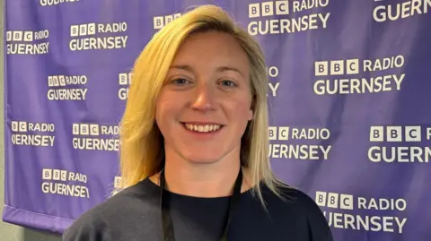 Charlie Cox is smiling at the camera as she stands next to a BBC Radio Guernsey branded banner. She has shoulder-length blonde hair and blue eyes., She is wearing a black top and a lanyard.