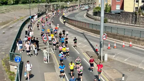 A group of people is running on a road next to a river. Some people are watching from the side of the road. The scene is seen from above.