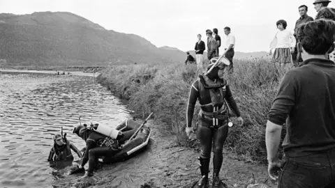 Mirrorpix via Getty Images Rescue divers at the edge of the Mawddach estuary in July 1966, watched by onlookers, during the search for survivors after the Prince of Wales pleasure boat sank. The divers are in full wetsuits and have scuba diving equipement, air tanks, masks and snorkels. 