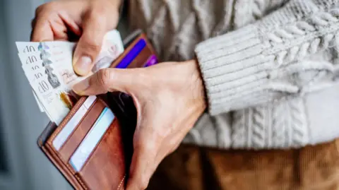 A man holding a brown wallet and notes in his hand. He is wearing a grey jumper. 