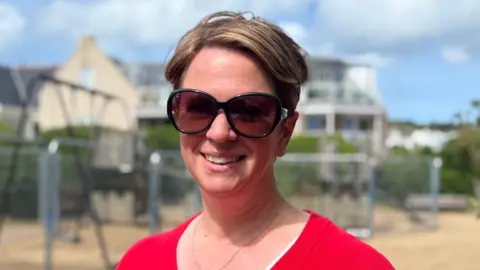 A woman called Rebekah Key smiles while standing in a play park. She has shirt fair hair. She has large sunglasses on and a red jumper.
