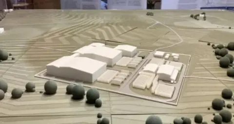 Guy Campbell/BBC A model of energy infrastructure. The model, on a large tabletop, shows white buildings within a large fenced compound, which is surrounded by fields.