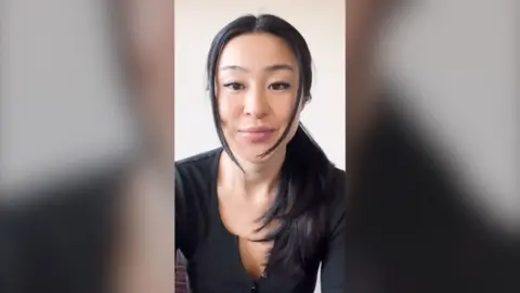 A woman with long black hair looks into the camera. She wears a black top