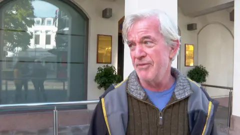 Gary Coats wearing a brown jumper with a zip in the middle with a dark and light grey coat. He is looking to the left and is stood in front of a white building. There is a large window on the left and two gold signs either side of a door. There are two plants under each sign. 