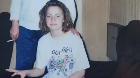 Family A teenage Katriona wears a white t-shirt with "Our Girls" written on it. An adult stands behind her in a white jumper and blue trousers carrying a cigarette. 